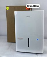 Front view of the white Midea dehumidifier showing logo, water level indicator, and the original packaging box behind it.