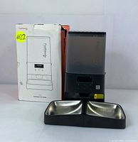 Photo showing the automatic pet feeder, two stainless steel bowls, and original box with some damage.