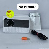 Front view of K8 mini projector with lens and speaker grille, power cable, user manual, and orange USB flash drive. Yellow sticker indicates price and note for no remote.