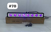 Front view of the LED wall effect light bar illuminated with purple light showing 8 beam lenses. Unit is placed on a cardboard box labelled 'LED WALL EFFECT LIGHT'. A plastic bag contains mounting accessories in front of the unit.