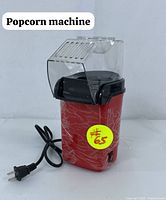 Front view of the red PM-1200 popcorn machine showing the transparent plastic top, power cord, and power switch on the side.