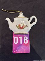 Front view of the teapot ornament showing the floral pattern and hanging string against a black background with inventory tag.