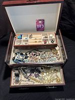 Open wooden jewelry box with two pull-out trays showing rings, bracelets, necklaces and other pieces