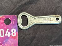 Photo of the Coca-Cola metal bottle opener showing the front with embossed Coca-Cola logo and bottle opening end.
