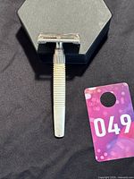 Top-down photo of vintage Gillette J-1 razor with ribbed handle on black surface with tag showing number 049