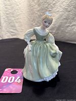 Front view of the vintage Royal Doulton bone china figurine showing the full figure in a pale green dress with puffed sleeves and blue bow in hair, standing on a flat black surface.