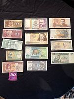 Wide view of 14 assorted world banknotes laid out on black background showing various historical figures, cultural symbols, and landscapes.