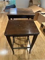 Two wooden nesting tables showing the tops and legs, placed on wooden floor with some boxes in background.