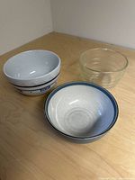 Three bowls shown together on wood surface: clear glass ribbed bowl, white ceramic bowl with blue inscription, and blue Denby bowl.