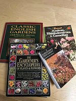 Four gardening books laid out on a wood surface showing covers: 'Classic English Gardens', 'Azaleas, Rhododendrons & Camellias' by Sunset Books, 'A Gardener's Testament', and 'The Gardener's Encyclopedia of Plants & Flowers'.