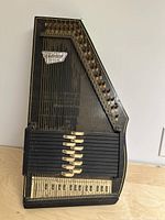 Photo showing full view of vintage black Oscar Schmidt Autoharp with numerous strings and chord bars, resting upright on wooden surface against wall.