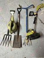 Full view of gardening tools including grass trimmer, small pitchfork, small shovel, digging tool, and watering can on floor.