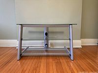 Front view of the TV stand showing metal frame, glass top and shelf, with central support and cable management holes.