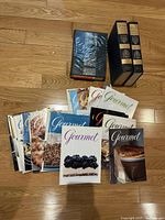 Photo of 23 total items including three hardcover cookbooks stacked and a spread of 20 Gourmet magazines fanned out on a hardwood floor.