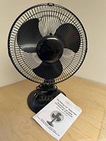 Black 12 inch Mainstays oscillating table fan with three blades on wooden surface with owner's manual placed in front.