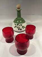 Photo of olive oil decanter with painted olive branches and three cranberry glass tumblers arranged around it on white background.