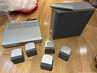 Sony DAV-SB100 DVD home theatre system with 5 silver satellite speakers, large black subwoofer, and silver DVD player unit on wooden floor.