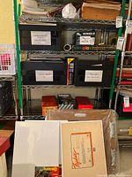 Shelving unit with labeled boxes containing various art papers and supplies including watercolor paper, litho paper, pastels, and acrylic paints, with canvases in front.