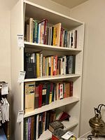 Front view of white manufactured wood bookcase filled with books and some miscellaneous items on five shelves.