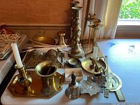 Overview of lot with brass candle sticks, mortar and pestle, metal hand bell, tray, and other brass items on table.