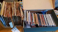 Box with large collection of vintage 45 RPM vinyl records in paper sleeves standing vertically next to an open box containing a series of CDs in jewel cases.