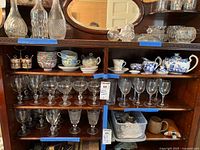 Wide view of wooden shelf holding various glassware and porcelain, including the blue and white Royal Crown Derby teapot, cream and sugar containers, and a floral cup and saucer with the noted chips.