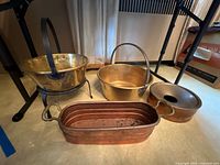 Four copper pots and planter arranged on floor with metal stands. Two pots have handles and there is one pot with a lid. The planter is rectangular with handles and visible dents.