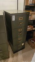 Photo showing olive green, four-drawer metal filing cabinet with brass handles, visible scratches and wear, positioned in basement setting