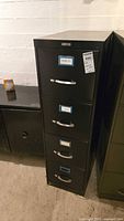 Black four drawer metal filing cabinet standing upright showing scratches and overall condition