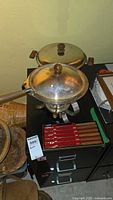 Westbend Discovery electric skillet with glass lid, metal fondue pot with wooden handle and glass lid, and set of fondue forks with red handles displayed on a black cabinet.