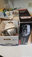 Kitchen items arranged in box including colanders, glass measuring cups, and strainers, alongside boxed roasting pan on countertop.