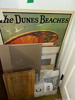 Vintage poster with 'The Dunes Beaches' text visible, assorted frames stacked in front, some glass panes and matting visible