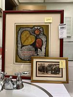 Framed 20x17 Leonard H. Mersk print with leaves centered, and smaller 7x9.5 framed print with park scene, displayed on a counter near sink faucets.