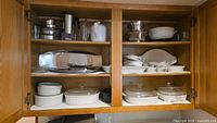 Full view of cabinet shelf showing metal serving trays, white casserole dishes, glass bakeware, metal and ceramic bowls, and white teapot