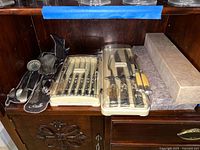 Wooden shelf surface showing metal cake cutter and server, metal strainer ladle, metal tongs, carving set with knife and fork in plastic case, and matching steak knives in plastic case.
