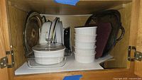 Overall cabinet shelf view showing multiple silver plated trays, stack of white ceramic ramekins, glass baking dish with lid, and additional white ceramic baking dishes.