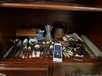 Wide shot of multiple items including metal spoons, coasters, strainer, grape shears, sugar tongs, and corkscrew on wooden shelf.
