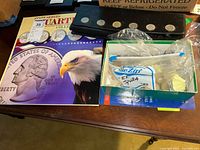 Photo of the collector board, folder with eagle and quarter image, plastic bag with extra quarters, and a box with mixed coins on a wooden table.