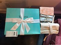 Tiffany box tied with white satin ribbon bow with three folded fabric travel pouches in peach, light blue, and off-white colors beside it.