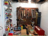 Wide angle photo showing pegboard with various hand tools hanging including saws, hammers, brushes, utility knives, manual drill, along with shelving containing paint and adhesive containers, spray oils, storage boxes, and small hardware containers in a basement workshop.