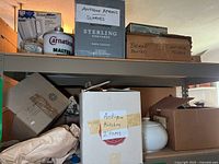 Shelf holding Carnation malted milk jar, boxes labeled antique aprons, broken painted plates, and antique kitchen items including a white ceramic sugar bowl.
