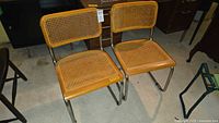 Full view of both vintage metal framed chairs showing cane seats and backs with tubular metal support frames.