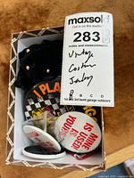 Top-down photo of cardboard box with black clip-on earrings and multiple pins including 'MINE IS USED! IS YOURS?', 'PLAY SARGON TOO', and an info card titled 'The Penny Pin'