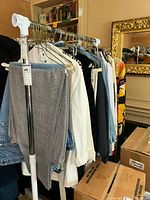 Side view of hanging vintage ladies clothing including pants, jeans, shirts, and dresses on a rack indoors near boxes.