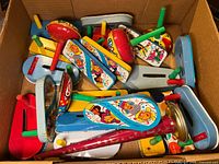 Top-down view of the lot showing multiple tin noise makers with colorful cartoon designs and wooden handles in a cardboard box.