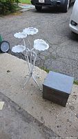 Photo showing white enameled multi-stemmed metal plant stand next to the metal Ware Dairy milk delivery box on pavement outdoors.