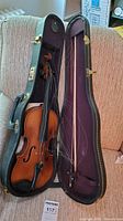 Vintage violin in open black case with purple interior on a beige couch, visible loose or broken strings and chin rest, shot from a distance.