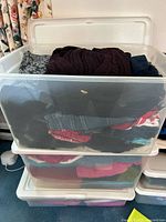Three stacked plastic bins containing assorted women's clothing including sweaters, jeans, and casual tops folded inside.