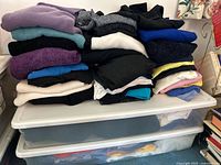 Stack of folded women's sweaters in various colors on a table with two clear storage bins underneath containing t-shirts.