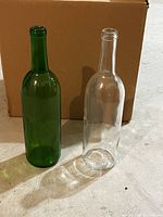 One green and one clear wine bottle standing upright in front of a cardboard box.